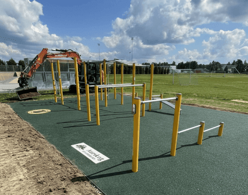Lauko treniruoklių zona su žalia gumine danga sporto aikštyne, šalia matoma statybinė technika ir futbolo stadionas